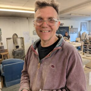Henry smiling at a camera in the Gray's Stone Carving workshop. He is wearing warm layers that have stone carving dust settled in the creases