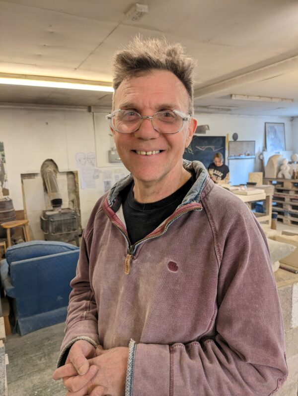 Henry smiling at a camera in the Gray's Stone Carving workshop. He is wearing warm layers that have stone carving dust settled in the creases